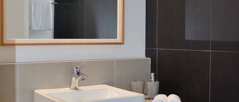 Une salle de bain moderne avec un grand miroir, un lavabo blanc et des murs recouverts de carreaux foncés.