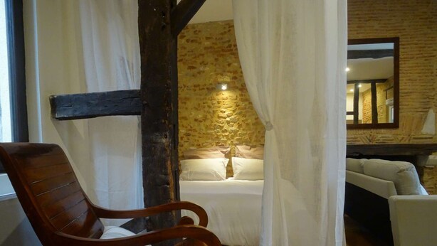 Une chambre confortable avec une chaise en bois, un lit recouvert de draps blancs et un mur en pierre.