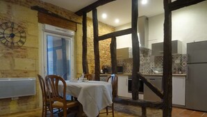 Un coin repas avec une table dressée pour deux personnes, des chaises en bois, une horloge au mur et une cuisine en arrière-plan.