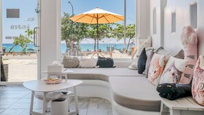 A modern interior with a large window overlooking a beach, a white sofa with cat-themed cushions, and a small table with a basket. - Cat at Greece (Hengchun)