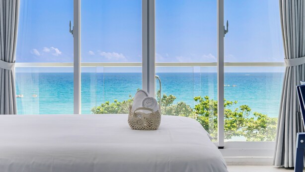 A bedroom with a large window overlooking the ocean, a white bed, and a decorative basket on the bed. - Cat at Greece (Hengchun)