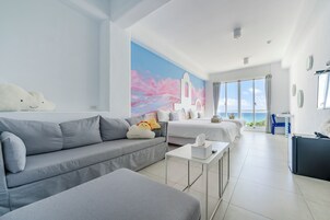 Una habitación de hotel moderna con un sofá gris, una mesita, una cama con ropa de cama blanca y un ventanal con vistas al mar.