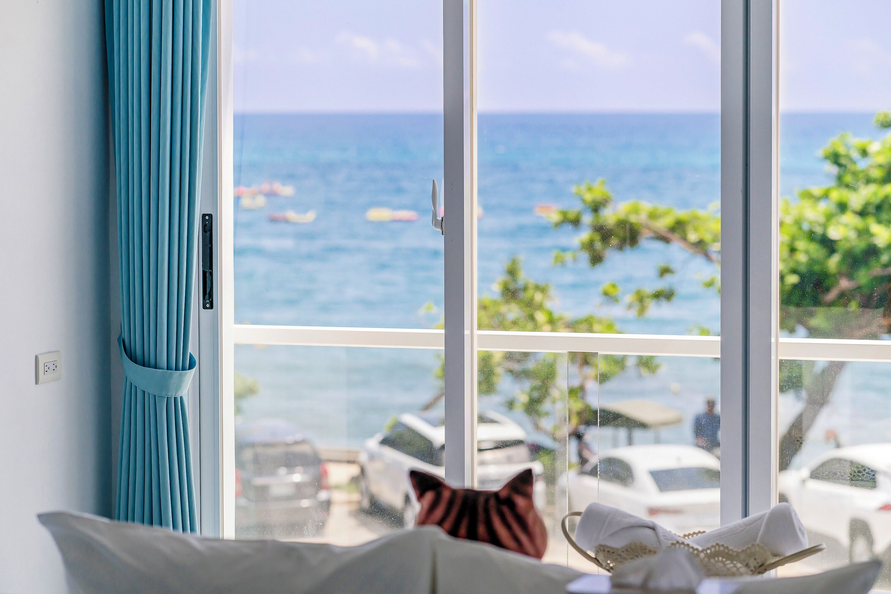 A room with a large window overlooking a beach, a blue curtain, and a view of parked cars and a person walking outside.