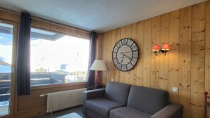 A room with a wooden wall, a grey sofa, a white table, a wall clock, and two wall-mounted lamps. - Cozy studio with Wifi near slopes in Tignes (Tignes)