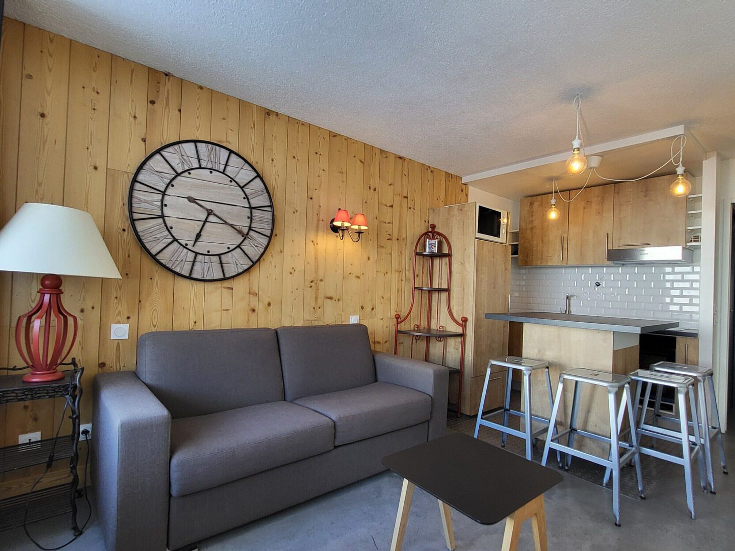 A modern living room with a grey sofa, a round wall clock, a red lamp, and a small table.