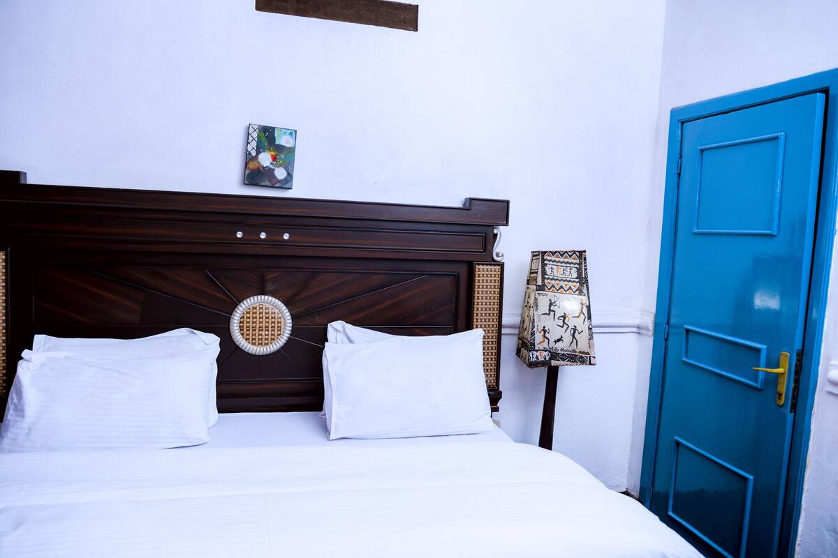 Un lit bien fait, avec du linge de lit blanc, une tête de lit en bois ornée d’un motif décoratif et une porte bleue sur le côté droit.