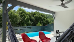Ein überdachter Außenbereich mit Pool, zwei roten Stühlen, einer Hängematte und einem Deckenventilator.