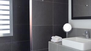 Ein modernes Badezimmer mit dunklen Fliesen, einem weißen Waschbecken, einem Spiegel und einer Holzbank.