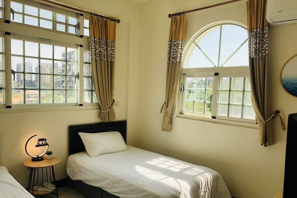 A bedroom with two beds, a bedside table with a lamp, and a large window with curtains.