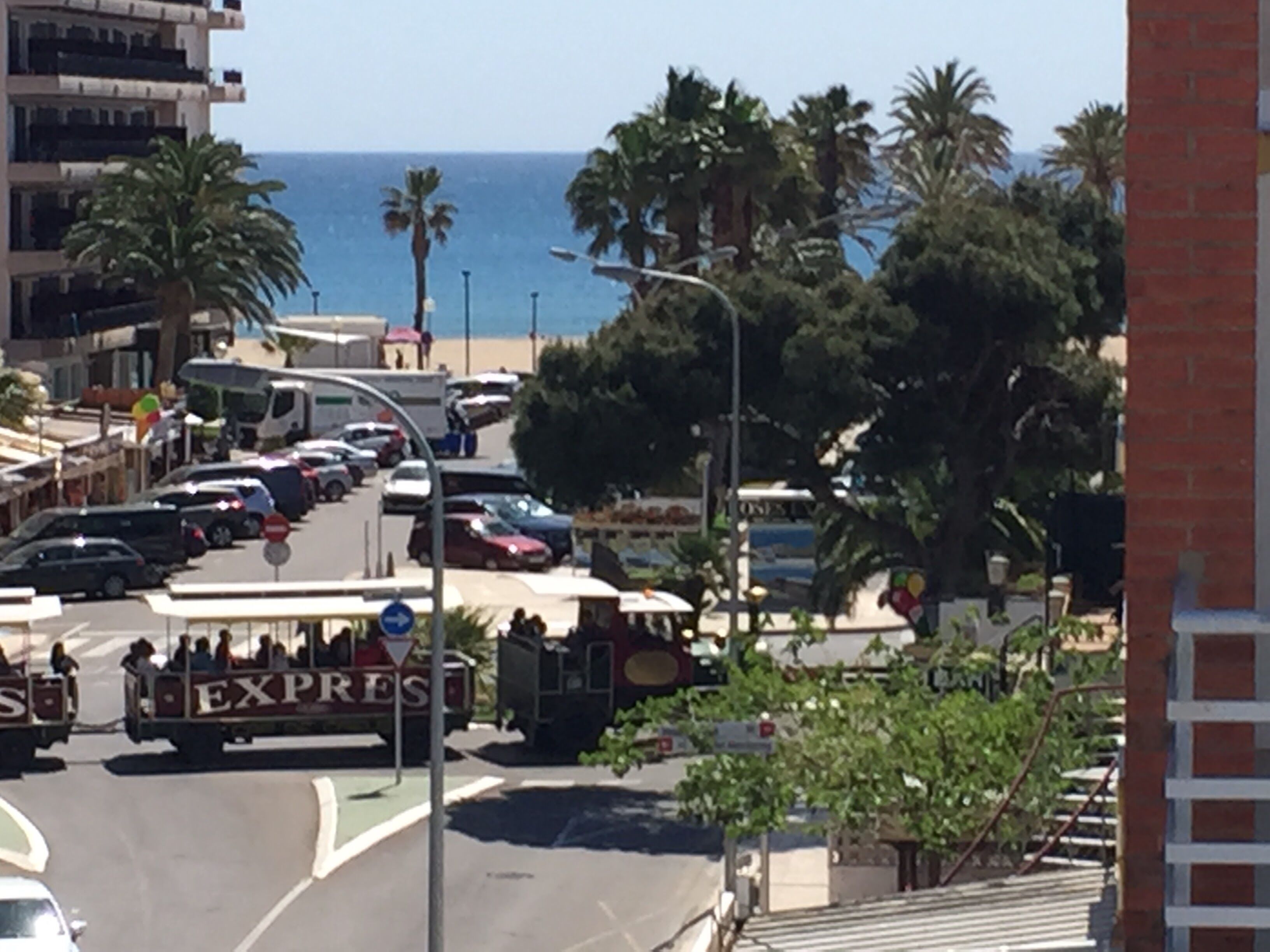 A coastal street with parked cars, a tram, and a view of the sea.