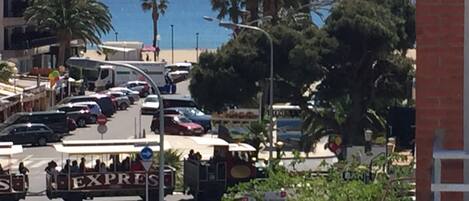 A coastal street with parked cars, a tram, and a view of the sea.