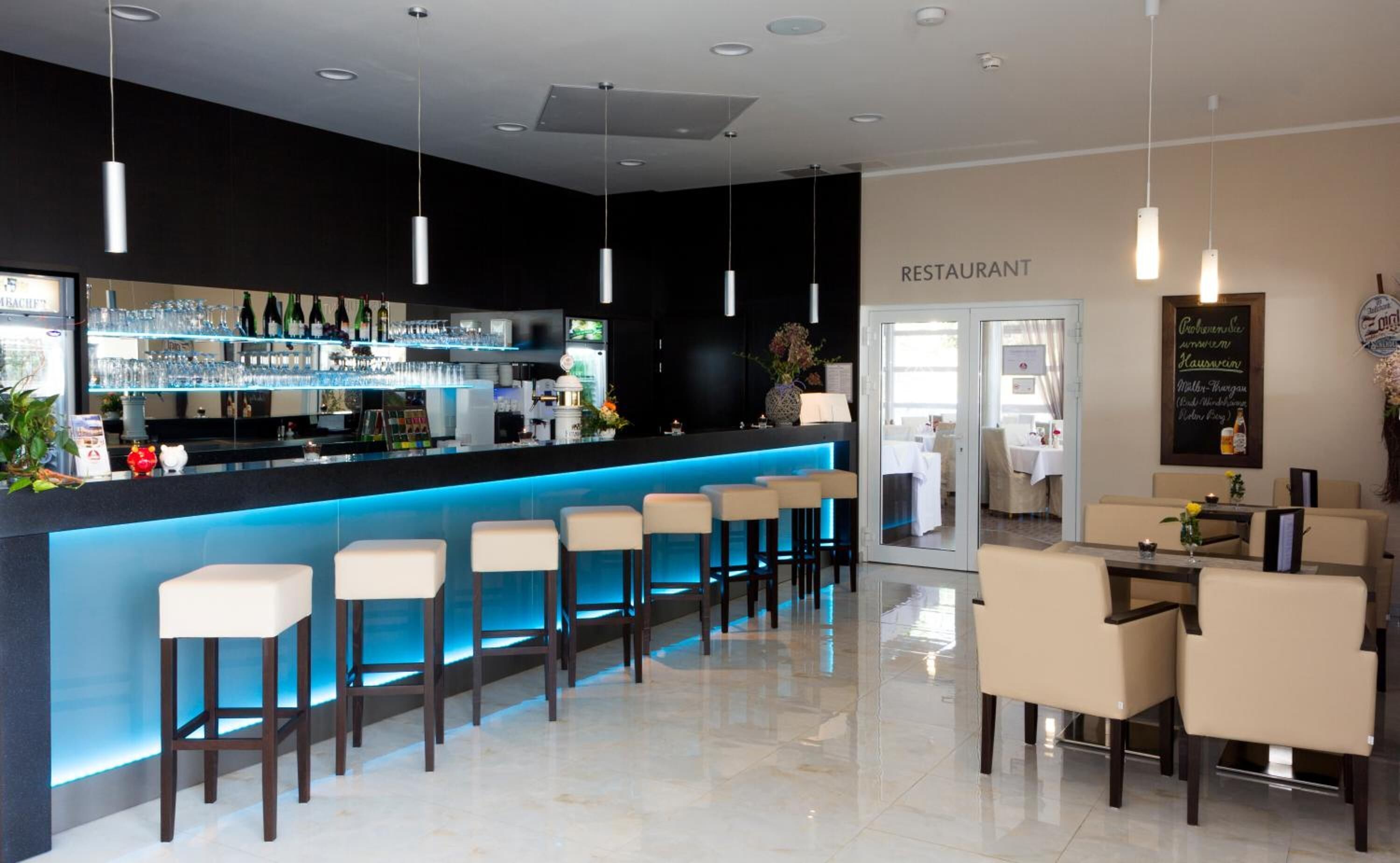 Zona de bar moderna con barra iluminada en azul, varios taburetes y vistas al área del restaurante.