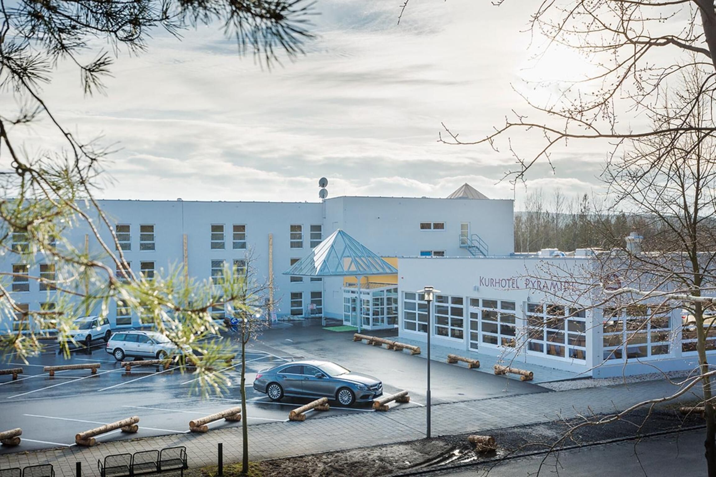 Et hotel med moderne design, med en markant glasfacade og en parkeringsplads med flere biler.