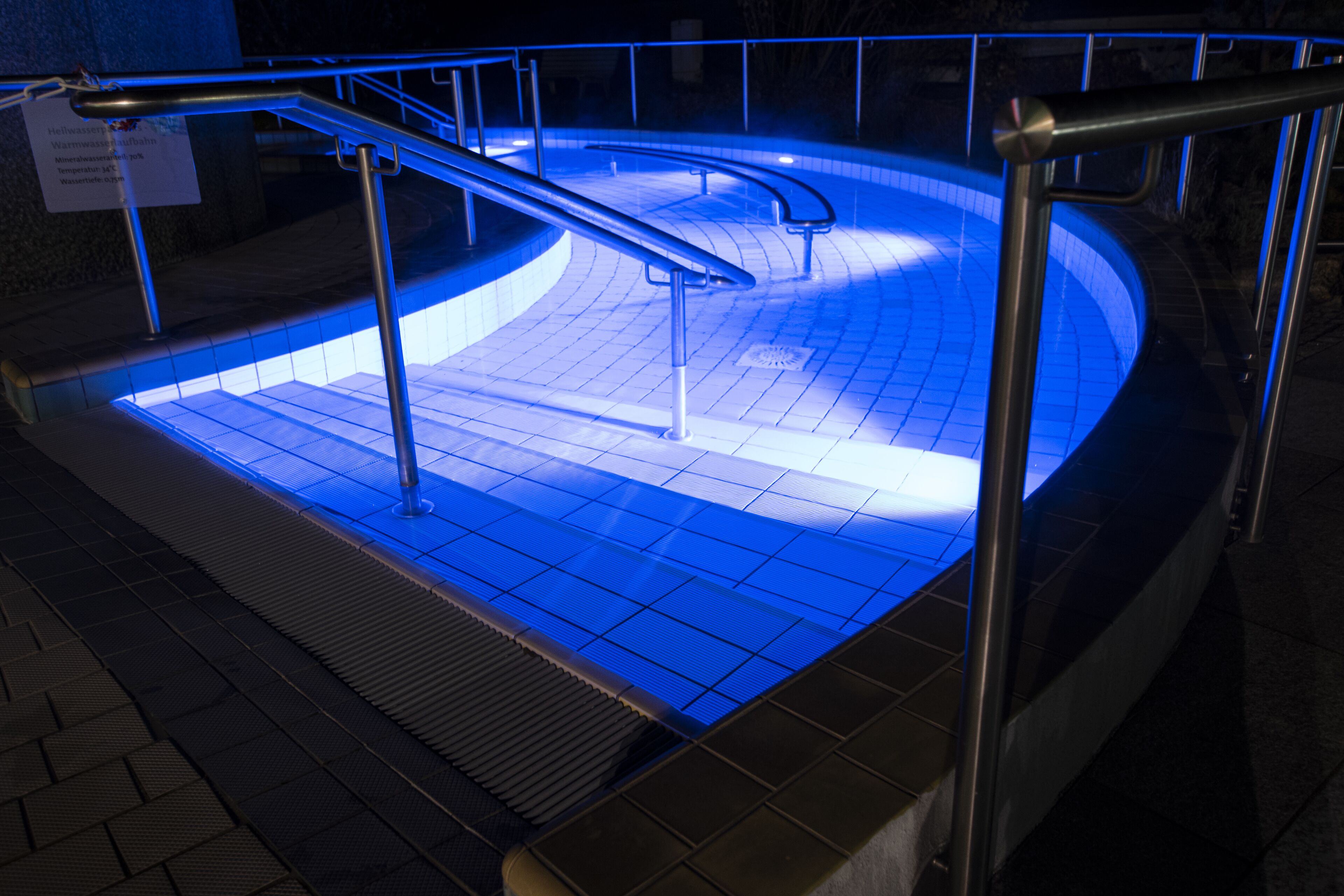 A pool area with a blue-lit floor, metal railings, and a sign indicating the pool's name and location.