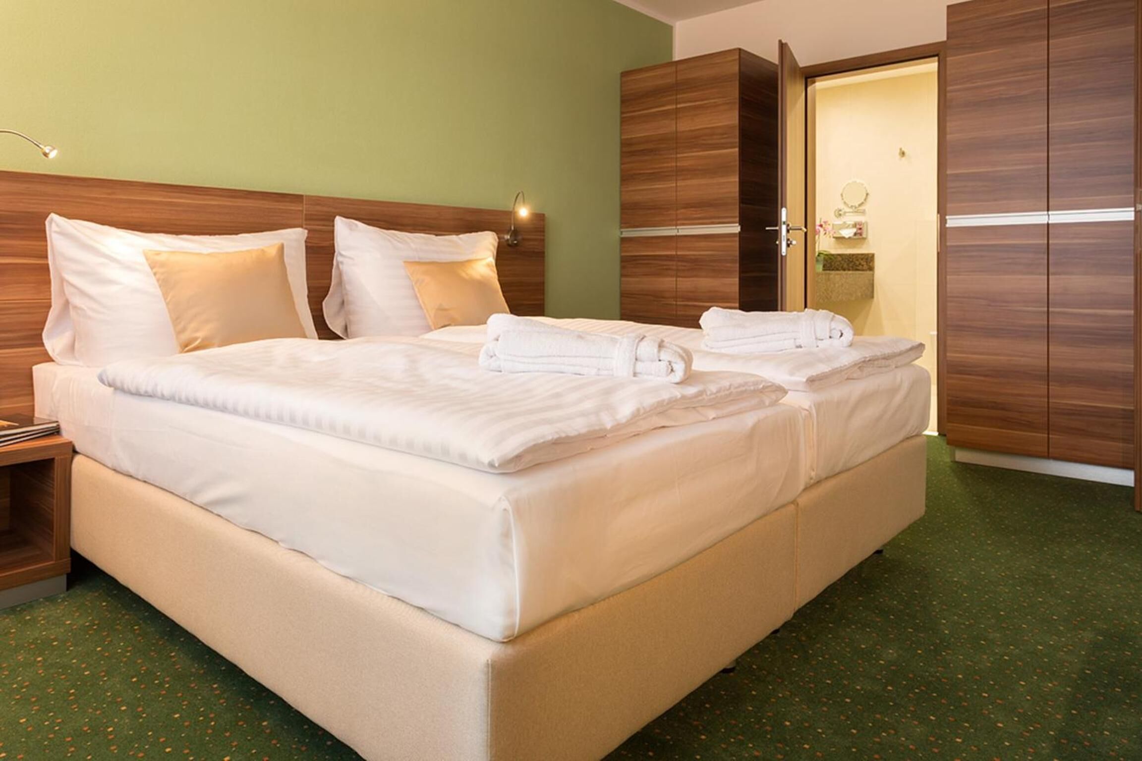 Una cama doble con ropa de cama blanca, una mesita de noche, un armario de madera y un piso cubierto de alfombra verde.