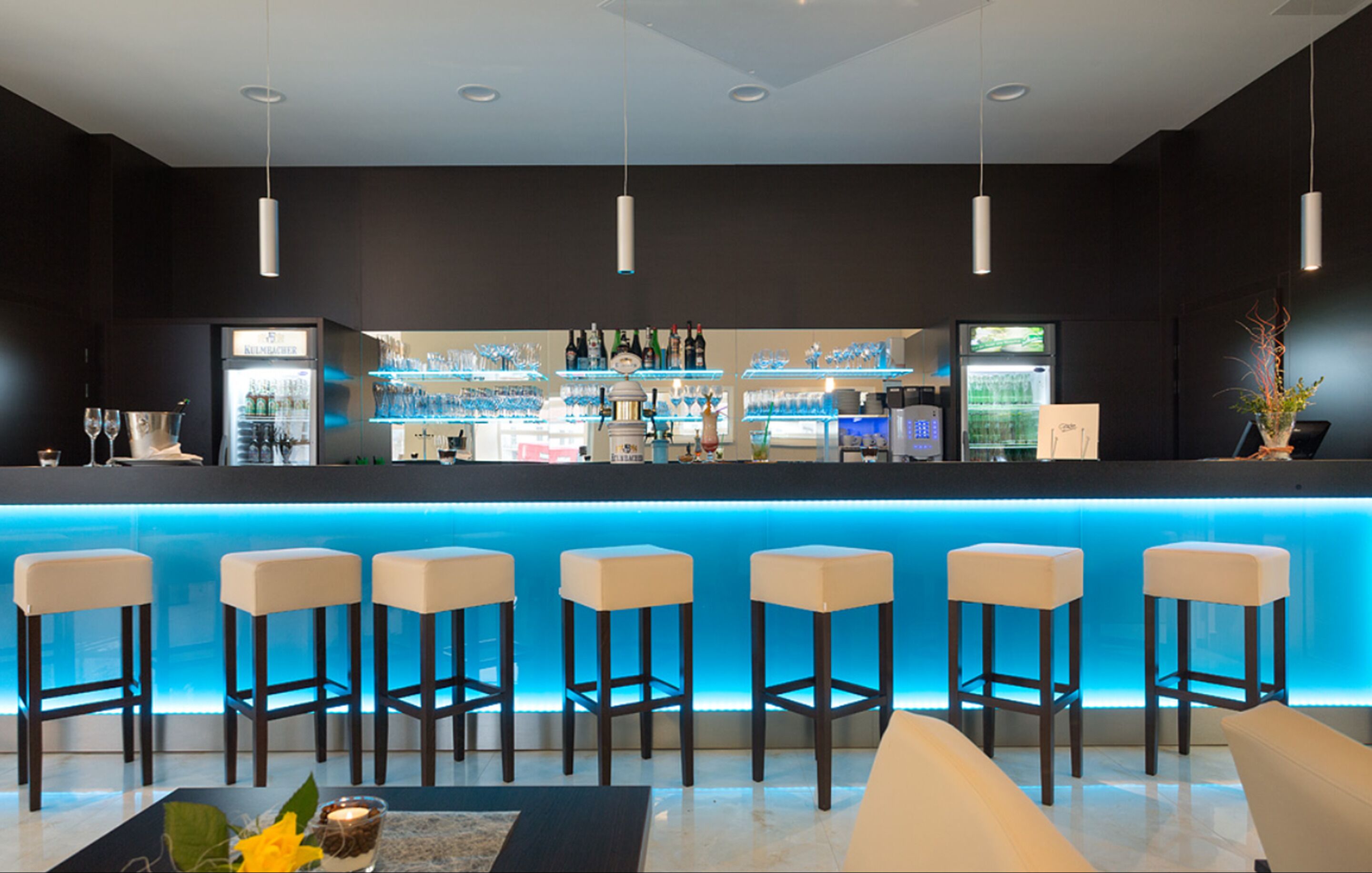 Eine moderne Bar mit einer blauen beleuchteten Theke, mehreren Barhocker und einer Auswahl an Flaschen und Gläsern.
