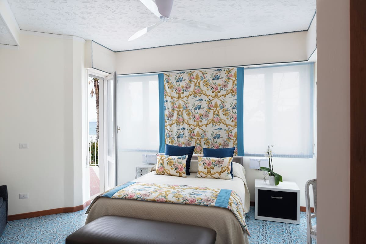 A bedroom with a bed, a nightstand, a plant, and a window with curtains.