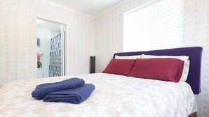 Een net opgemaakt bed met een bloemenprintdeken en twee kussens, één rood en één wit, in een kamer met behang met patroon en een raam met jaloezieën.