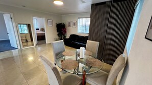 Een moderne woonkamer met een glazen eettafel, comfortabele zithoek, een zwarte leren bank en een groot raam met zonwering.