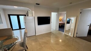 Een ruime woonkamer met een flatscreen televisie aan de muur, een glazen eettafel en een deur naar een slaapkamer.