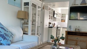 Sala de estar com um sofá branco, uma mesa de centro de vime e uma cozinha ao fundo.