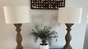 Uma mesa de madeira branca com duas lâmpadas, um vaso de planta e um expositor de conchas na parede.