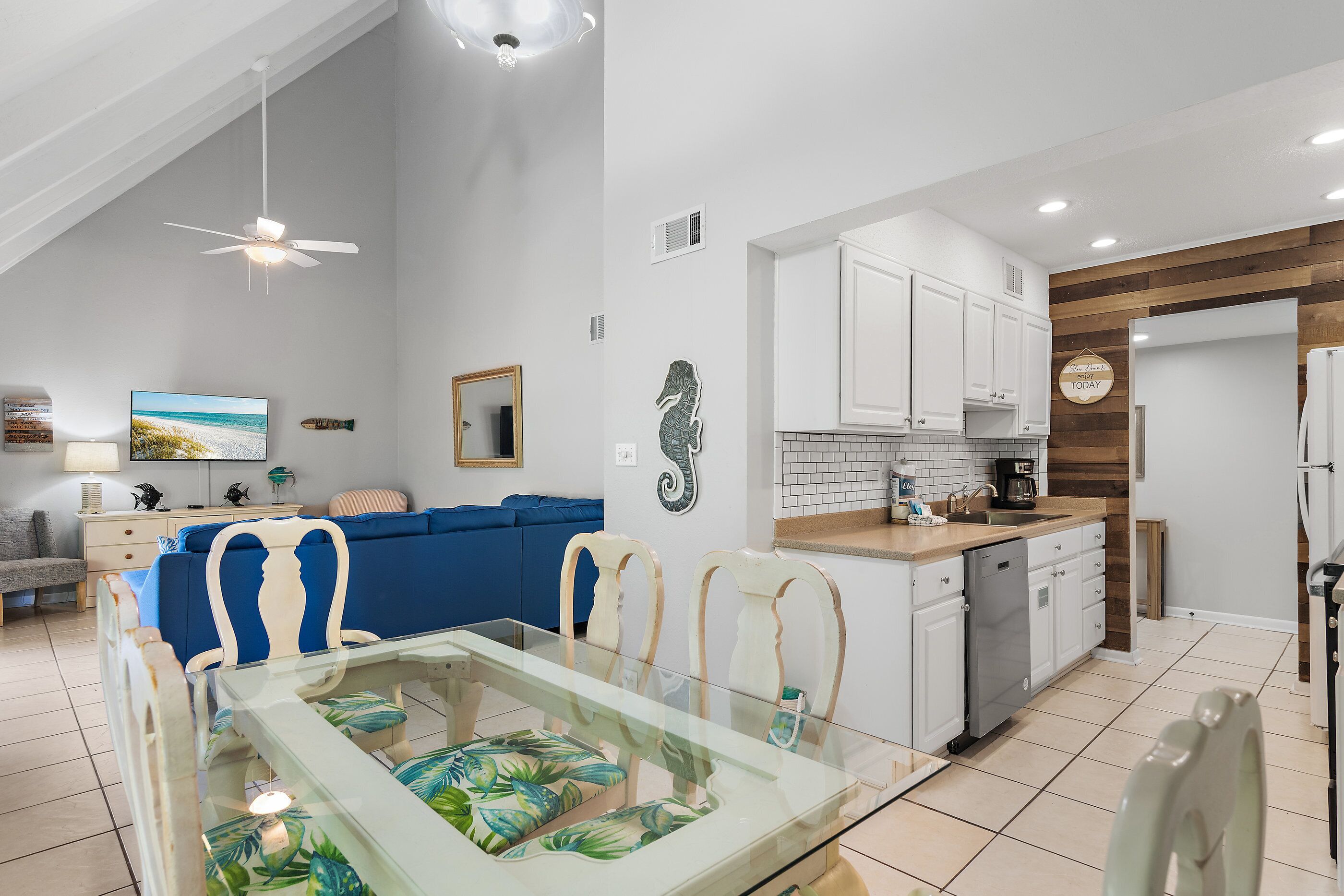 A modern kitchen with white cabinets, a dining area with a glass table, and a living room with a blue sofa.
