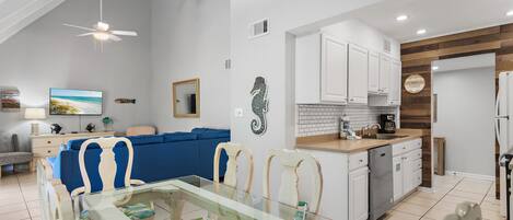 A modern kitchen with white cabinets, a dining area with a glass table, and a living room with a blue sofa.