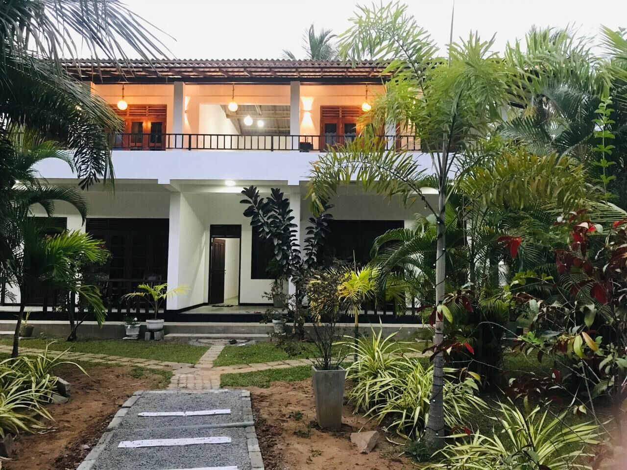 Un edificio de dos pisos con balcón, rodeado por un jardín con diversas plantas y un sendero pavimentado que conduce a la entrada.