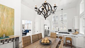 Een ruime keuken met witte kasten, een houten eettafel met rieten stoelen en een grote kroonluchter.