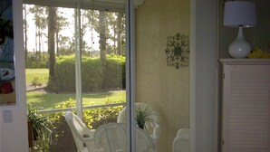 A room with a glass door leading to a balcony with white plastic chairs and a table. There is a white cabinet with a lamp on top and a wall decoration. - 2 Bed 2 Bath First Floor Condo in Stoneybrook of Estero Golf Community  (Estero)