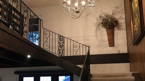 Una escalera con barandilla de madera y peldaños alfombrados que conducen a un rellano. Una barandilla metálica decorativa con diseños intrincados bordea la escalera. Una lámpara colgante con múltiples bombillas está suspendida del techo. Una planta en maceta está colocada en la pared del rellano.