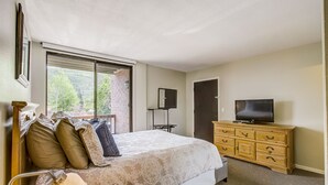 A bedroom with a large bed, a dresser, a television, and a window with a view of trees.
