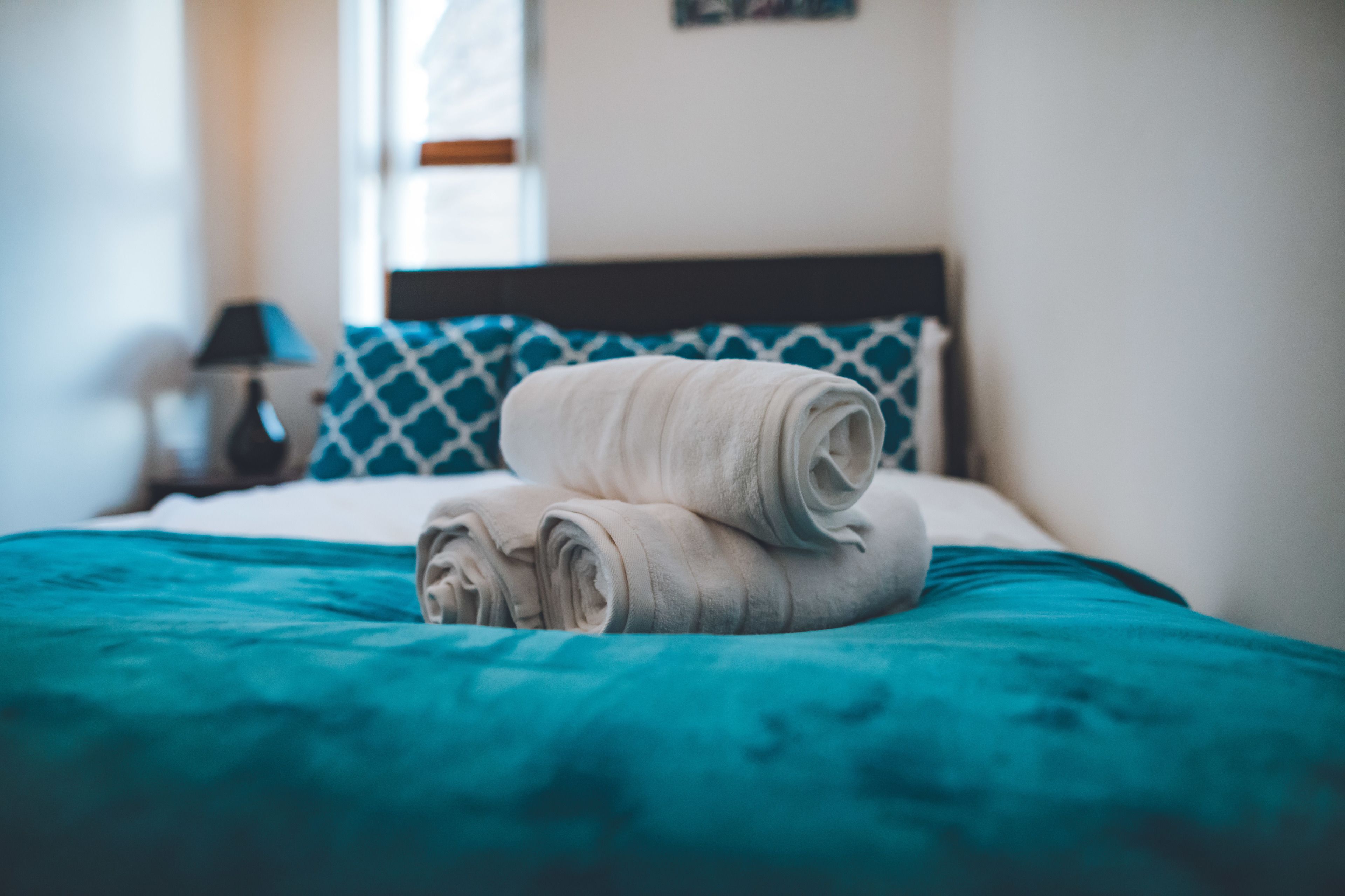 A neatly made bed with a teal bedspread and two rolled towels.