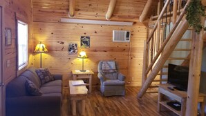Interior acollidor d'una cabana amb sòl de fusta, un sofà, una cadira, una taula petita, un televisor sobre un suport de fusta i una escala.