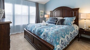 A bedroom with a large bed, a wooden headboard, a ceiling fan, a nightstand with a lamp, and a window with sheer curtains.