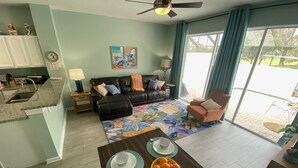 A modern living room with a black leather sofa, a colorful floral rug, and a dining area with a wooden table set for four.