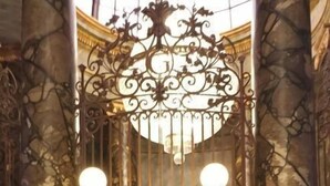 A person seated at a podium in a room with ornate columns and a chandelier.