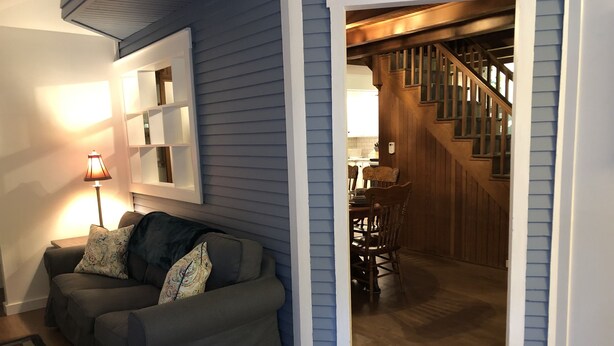 A living room with a gray sofa, patterned cushions, a wooden staircase, and a dining area in the background.