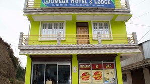 Un edificio de dos pisos con un letrero que dice Omega Hotel & Lodge. Tiene un restaurante y bar en la planta baja, con macetas de plantas al frente.