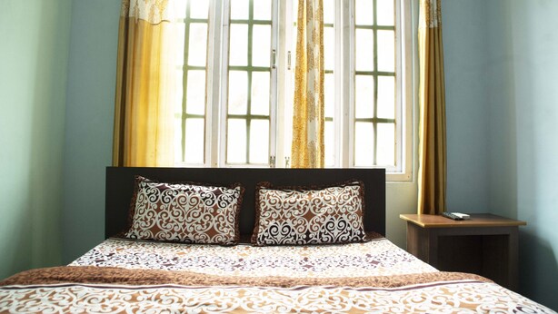 Un dormitorio con una cama, dos almohadones estampados, una mesita de noche y una ventana con cortinas.