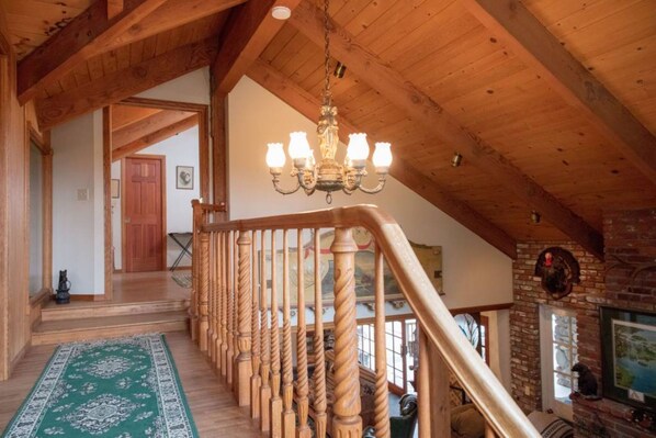 Un escalier en bois surmonté d’un lustre, un tapis à motifs verts et une cheminée en briques.