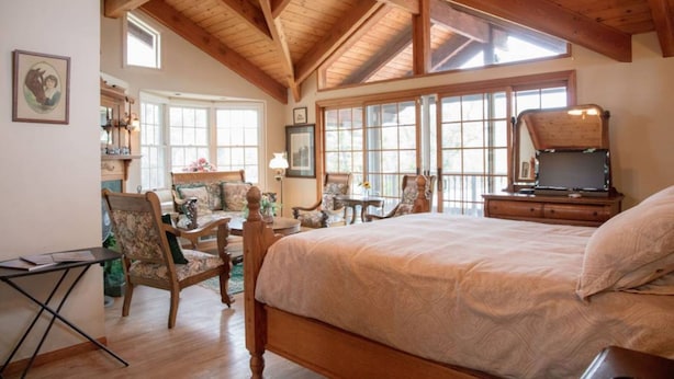 Une chambre spacieuse dotée d’un grand lit, de poutres apparentes en bois au plafond et de nombreuses fenêtres qui laissent entrer la lumière naturelle.