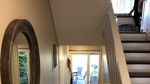A hallway with a wooden floor, a mirror, a lamp, and a staircase leading to a balcony with a chair and table. - 3 Bedroom, 3 Bath Oceanside Beach Villa (Hilton Head Island)