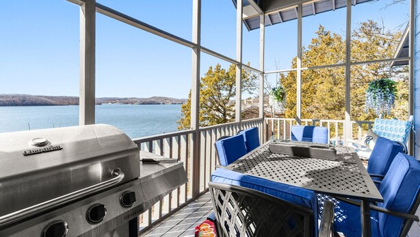 A covered outdoor deck with a grill, blue cushioned seating, and a view of a lake and trees. - Margaritaville Resort~ Close to the Pool~ Large Home-Amazing Water Views-Recently Redecorated (Osage Beach)