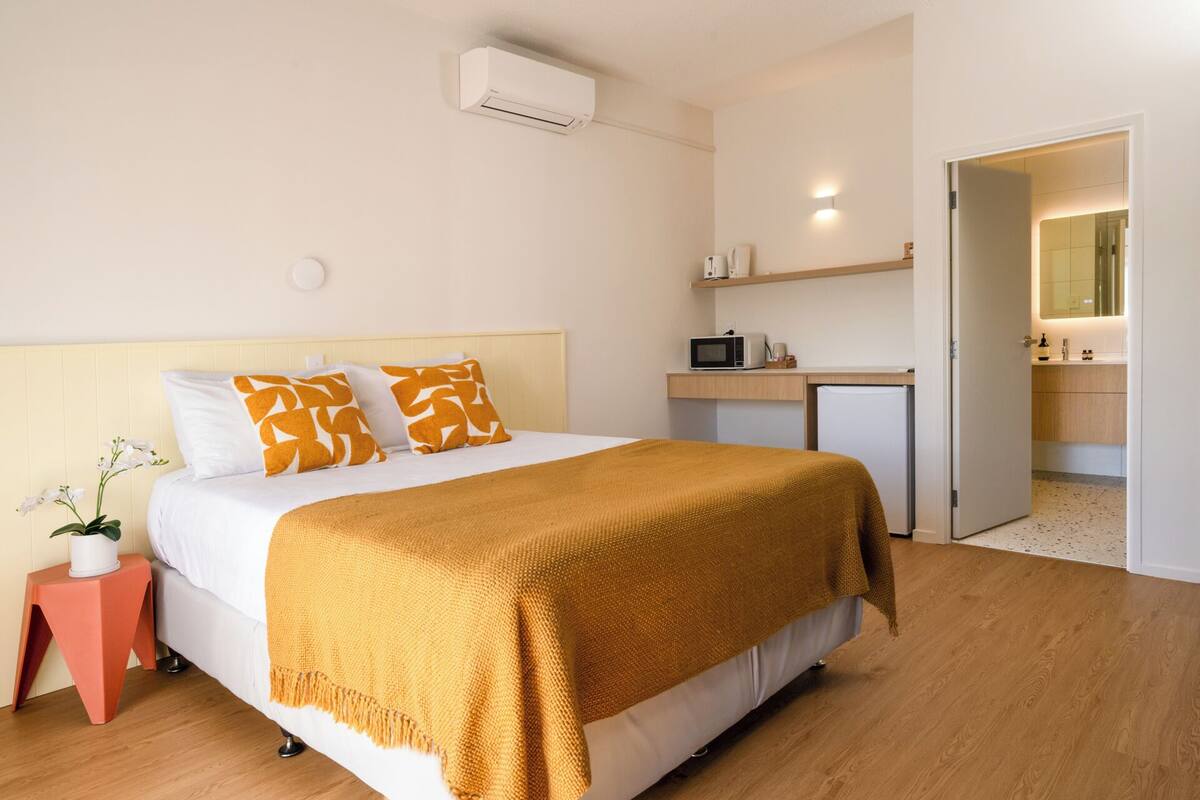 A bedroom with a bed, a bedside table with a potted plant, a microwave, and a small kitchenette.