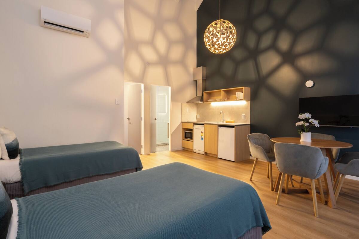 A modern room with two beds, a kitchenette, a dining area, and a ceiling light fixture.
