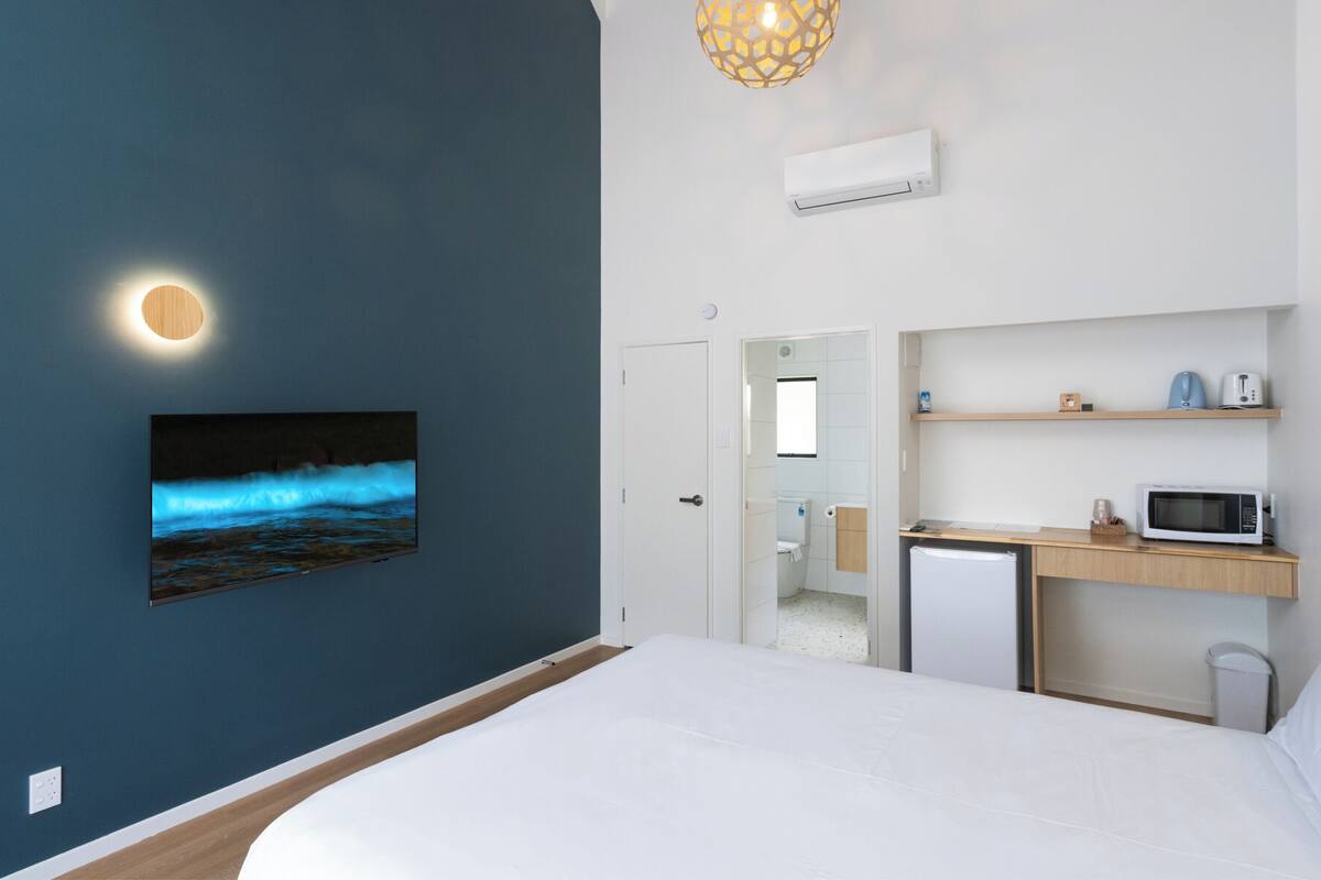 A modern bedroom with a bed, a wall-mounted TV displaying a seascape, a small kitchenette with a microwave, and a ceiling light fixture.