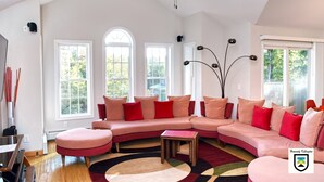 A spacious living room with a large pink sectional sofa, a ceiling fan, and multiple windows providing natural light.