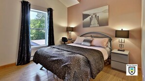 A bedroom with a large bed, two bedside lamps, a nightstand, a window with curtains, and a painting on the wall.